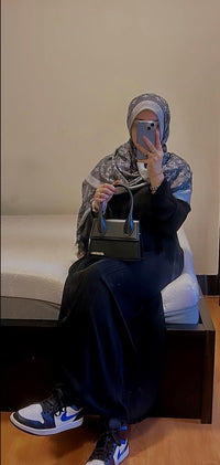Woman in black abaya and printed hijab sitting on bed, holding black handbag, wearing sneakers