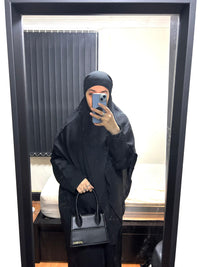 Woman in black open abaya taking mirror selfie, holding black handbag in bedroom