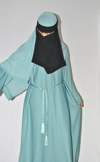 Light blue closed abaya with a braided belt and black niqab on a plain background