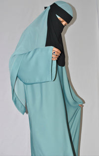 Woman in teal closed abaya with wide sleeves, wearing a black niqab on a plain background.