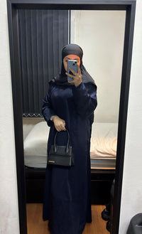 Woman wearing a navy blue abaya with black hijab, holding a black handbag, standing indoors
