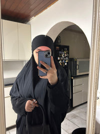 Woman in black abaya and hijab taking mirror selfie in kitchen holding black handbag