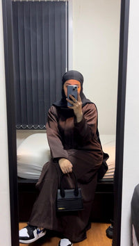 Woman in brown open abaya and black hijab taking a mirror selfie, holding black handbag