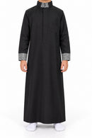 Boy wearing a black Saudi thobe with black and white patterned collar and cuffs, front view