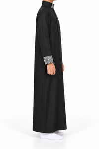 Side view of a boy wearing a black Saudi thobe with black and white patterned collar and cuffs