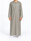 Boy wearing a cement grey Omani thobe with curved chest stitching, front view